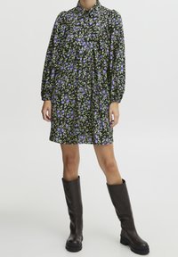 Blommig klänning i svart med lila och gröna mönster, har krage och långa ärmar, tillsammans med knähöga bruna stövlar.