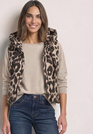 Mujer sonriente con cabello hasta los hombros que lleva un suéter beige, chaleco acolchado con estampado de leopardo y vaqueros azules sobre un fondo liso.