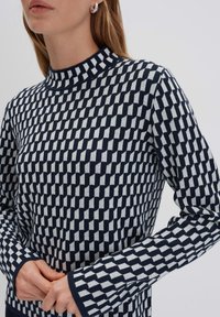 Marineblauer und weiß gemusterter Pullover mit hohem Kragen, der ein geometrisches Karomuster aufweist und lange Ärmel mit kontrastierenden Bündchen hat.