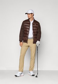 Smeđa puf jakna s rebrastim rukavima preko bijelog polo majice, bež hlača i bijelih golfštuća, drži golf palicu. Bijela kapa zaokružuje izgled.