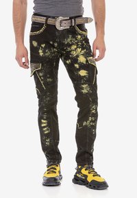 Schwarze Denim-Cargo-Hosen mit gelben Batikakzenten, genoppten Taschen und Strassdetails. Kombiniert mit gelb-schwarzen Sportschuhen.