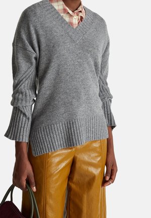 Personne portant un pull gris à col en V par-dessus une chemise à carreaux, un pantalon en cuir couleur moutarde, tenant un sac sombre par la poignée.