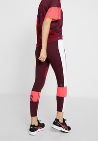Viininpunaiset urheiluleggingsit, joissa on valkoisia ja vaaleanpunaisia yksityiskohtia, valmistettu joustavasta materiaalista, yhdistetty viininpunaiseen ja vaaleanpunaiseen verkkopaitaan. Mustat lenkkarit.