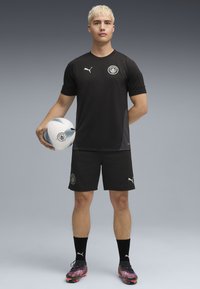 Un jeune homme en tenue de sport noire tient un ballon de football, se tenant fièrement contre un fond dégradé gris.