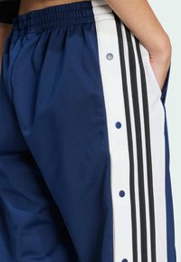 Person wearing navy blue track pants with white side panels, black stripes, and snap buttons, hand resting in pocket.