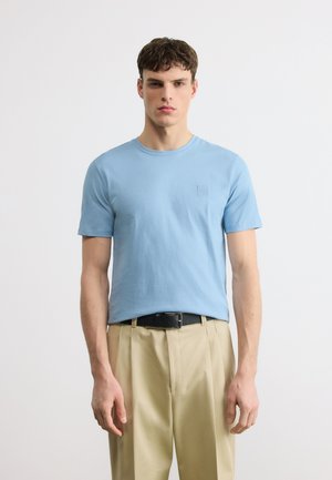 Νεαρός άνδρας που στέκεται, φορώντας γαλάζια κοντομάνικη μπλούζα, μπλεγμένη σε μπεζ πλισέ παντελόνια με μαύρη ζώνη, με απλό λευκό φόντο.