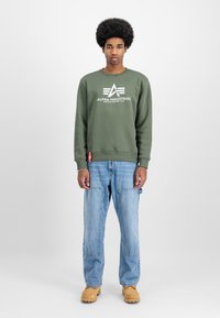 Ελιά πράσινη μπλούζα με λευκό λογότυπο, σε συνδυασμό με ανοιχτό μπλε φαρδιά τζιν και καφέ μπότες εργασίας. Απλός σχεδιασμός, χωρίς σχέδια.