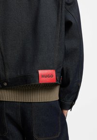 Dunkelblaue Jeansjacke mit einem gerippten olivgrünen Saum, rotem HUGO-Label und kontrastierenden Nähten. Verfügt über Druckknopfverschlüsse und seitliche Taschen.
