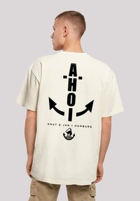 Crèmekleurig katoenen T-shirt met een zwart grafisch ontwerp op de achterkant, featuring het woord "AHOI," pijlen en een nautisch motief aan de onderkant.