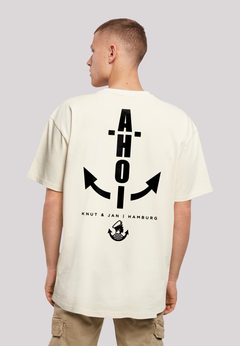 Crèmekleurig katoenen T-shirt met een zwart grafisch ontwerp op de achterkant, featuring het woord "AHOI," pijlen en een nautisch motief aan de onderkant.