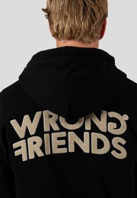 Černá mikina s kapucí, na zadní straně s velkým béžovým nápisem "WRONG FRIENDS". Materiál je měkký a strukturovaný, ideální pro volný čas.