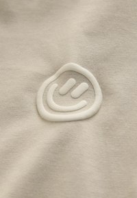 Hellbeiges Textil mit einem erhabenen weißen Smiley-Logo. Der Stoff hat eine glatte Textur mit subtilen Schattierungsvariationen.