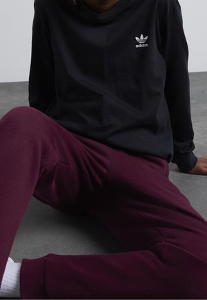 Persona sentada en el suelo con camiseta negra de manga larga de Adidas y pantalones burdeos con calcetines blancos sobre un fondo gris.