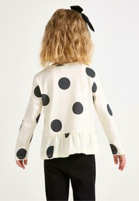 Langärmliges Shirt in Creme mit großen, schwarzen Punkten und einem rüschierten Saum. Hergestellt aus weichem Stoff mit lockerer Passform und rundem Ausschnitt.