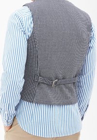 Šedá vesta s houndstooth vzorem a pruhovaným zadním dílem. Obsahuje detail s přezkou a páskem. Nošena přes modro-bílou pruhovanou košili.