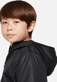 Veste à capuche noire en matériau lisse, dotée d'une fermeture éclair et d'épaules légèrement rembourrées, avec une coupe cintrée et sans motifs visibles.