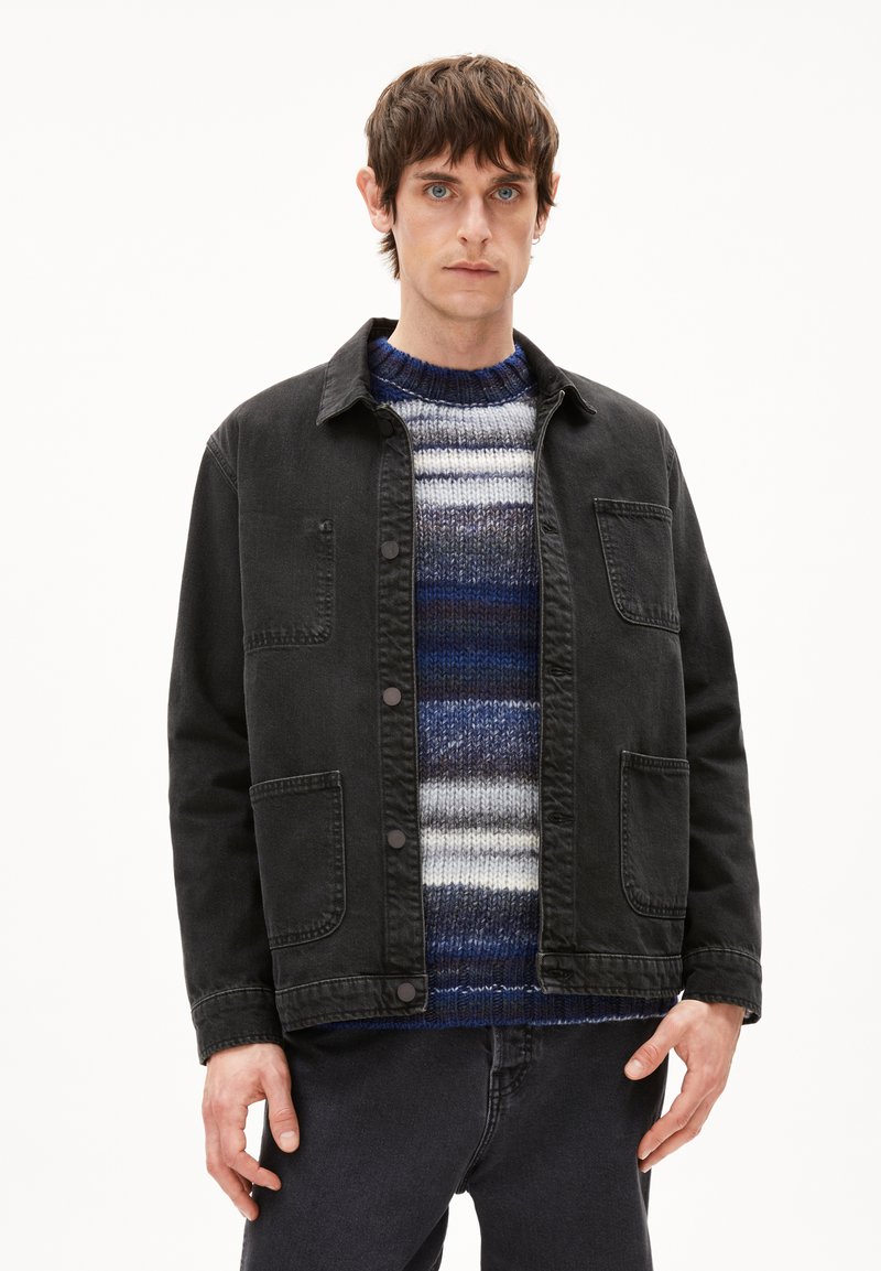 Schwarze Jeansjacke mit zwei Vorderentaschen, Knopfleiste und einem Kragen. Darunter ein blau-grau gestreifter Strickpullover.