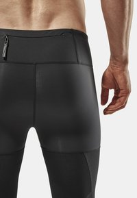 Leggings de sport noirs avec une texture lisse, dotés d'une poche zippée et de panneaux en mesh contrastants sur le bas des jambes.