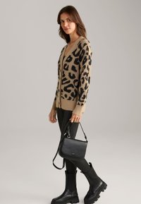 Cardigan beige con stampa leopardata nera, bordi a coste, abbinato a una borsa in pelle nera e stivali neri aderenti al ginocchio.