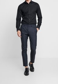 Czarna koszula na guziki z długimi rękawami, zestawiona z dopasowanymi ciemnymi spodniami w kratę i czarnymi skórzanymi butami. Dopasowany i elegancki fason.