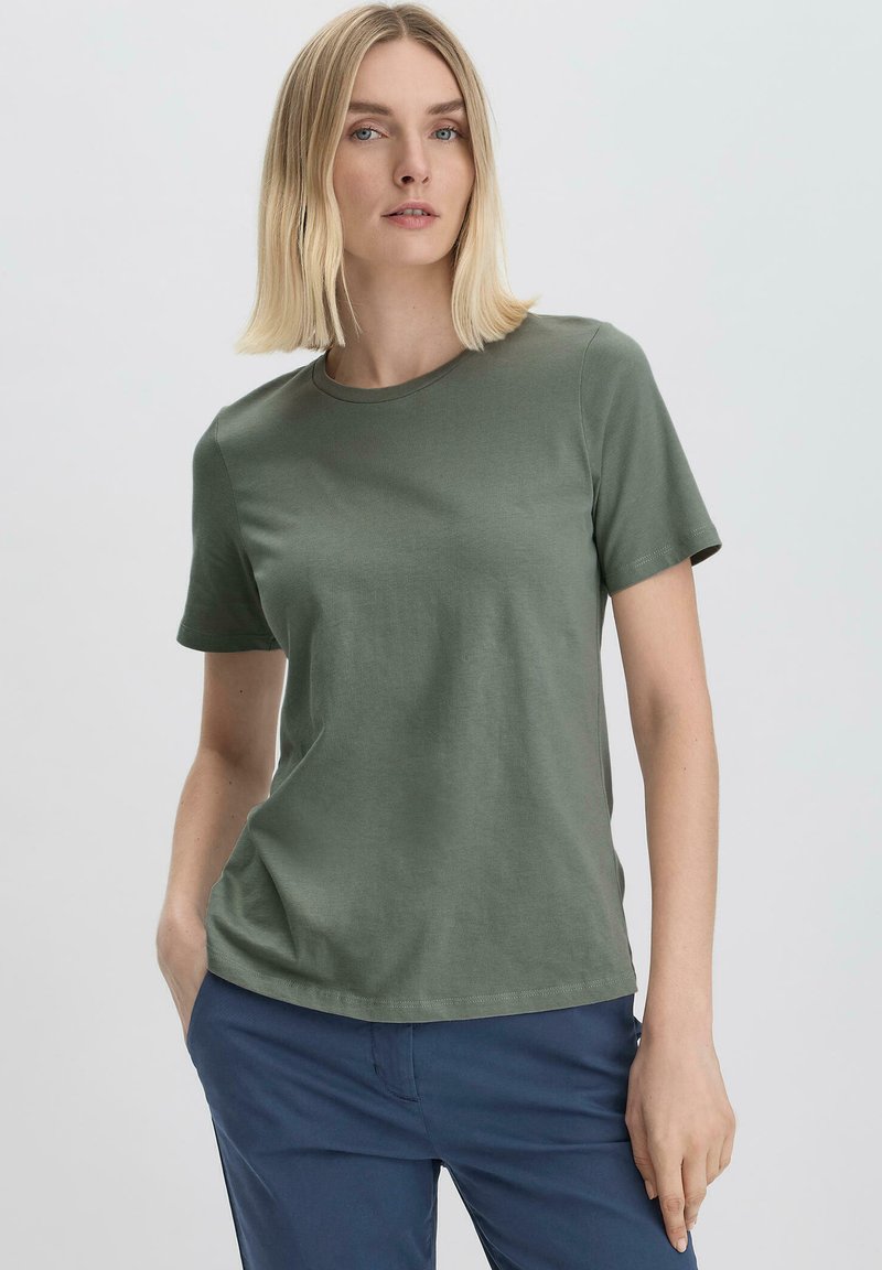 Vihreä puuvillapaita, jossa on pyöreä kaula-aukko ja lyhyet hihat. Sileä pinta, rento istuvuus, yhdistettynä sinisiin housuihin.