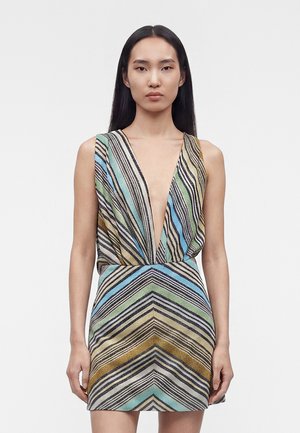 Kobieta o długich ciemnych włosach, ubrana w mini sukienkę bez rękawów z głębokim dekoltem w kształcie litery V, ozdobioną wielokolorowymi, ukośnymi i jodełkowatymi paskami.