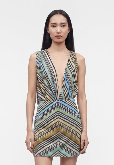 Femme aux longs cheveux foncés portant une mini-robe sans manches avec un décolleté en V profond et des rayures diagonales et chevrons multicolores.