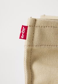 Sac en toile beige avec coutures renforcées et étiquette Levi's rouge attachée. Texture lisse sans motifs ni ornements visibles.