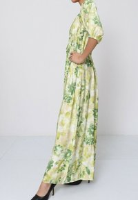 Robe maxi en tissu léger avec imprimé floral vert. Présente un buste smocké et des manches bouffantes, stylisée avec des talons hauts noirs.