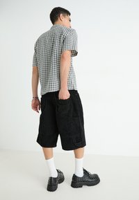 Ležérní outfit skládající se z černobílé kostičkované košile s krátkým rukávem, černých manšestrových šortek a robustních černých bot s hroty.