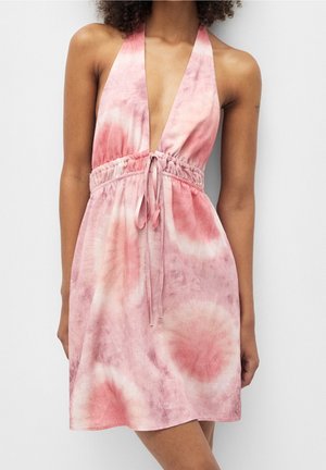 Femme portant une robe sans manches à col haut en tie-dye rose et blanc, avec un décolleté en V profond et une taille cintrée nouée avec un nœud.