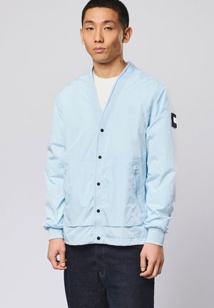 Jonge man die een lichtblauwe jas met knopen draagt, met een zwarte patch op de linkermouw en donkere broek, staand tegen een effen achtergrond.