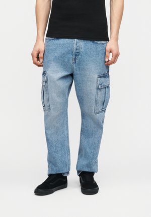 Personne portant un pantalon cargo en denim bleu clair avec des poches latérales, un t-shirt noir et des baskets noires, debout devant un fond uni.