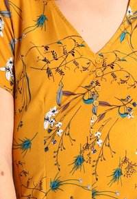 Sinappi keltainen pusero, jossa kukkakuvio, V-kaulus, lyhyet hihat ja viisi nappia edessä. Pehmeä, kevyt kangas.