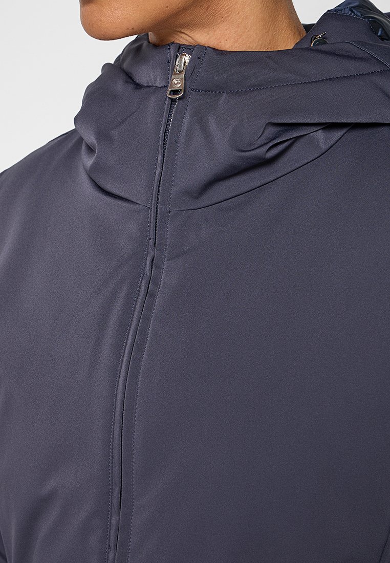 Närbild av en person som bär en mörkblå huvad jacka som är dragen upp till halsen, med en slät tyg och en silverdragning.