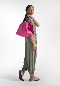 Bolso tote grande de color rosa con ribete negro y logo, hecho de material liso. La modelo lleva un conjunto suelto verde y sandalias rosa claro.