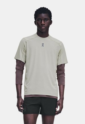 Jeune homme portant des chemises superposées beige à manches courtes et marron à manches longues avec un short noir, debout devant un fond uni.