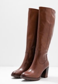 Bottes marron en cuir, montantes jusqu'au genou, avec un talon bloc haut, une texture lisse, une fermeture éclair sur le côté et une semelle renforcée pour une bonne adhérence.
