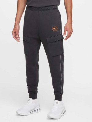 Joggers Nike de color gris oscuro con cinturilla elástica, bolsillos laterales y detalle del logotipo en color naranja; presentan un corte ajustado en la pierna y ribetes blancos en contraste.