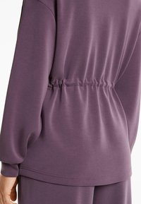 Lilla sweatshirt med indsnøret talje. Glat stof, lange ærmer og en afslappet pasform. Har en lige kant og synlige sømdetaljer.