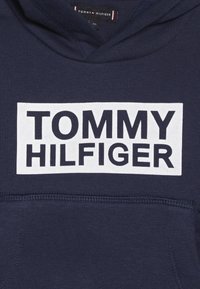 Granatowy hoodie z dużym białym prostokątnym logo, na którym znajduje się "TOMMY HILFIGER" w grubych czarnych literach. Wykonany z bawełny, z kieszenią typu kangur.