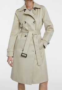 Abrigo trench beige de tejido suave, diseño de doble botonadura con botones grandes, cintura ceñida por un cinturón y bolsillos laterales.