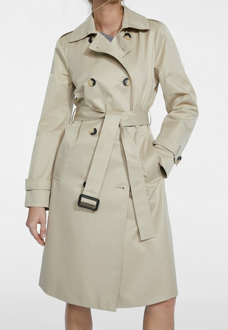 Abrigo trench beige de tejido suave, diseño de doble botonadura con botones grandes, cintura ceñida por un cinturón y bolsillos laterales.