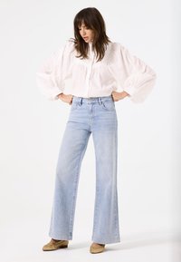 Blusa blanca de manga abullonada con detalles de encaje combinada con jeans de mezclilla azul de cintura alta y patas de elefante. Zapatos de mimbre color beige completan el atuendo.