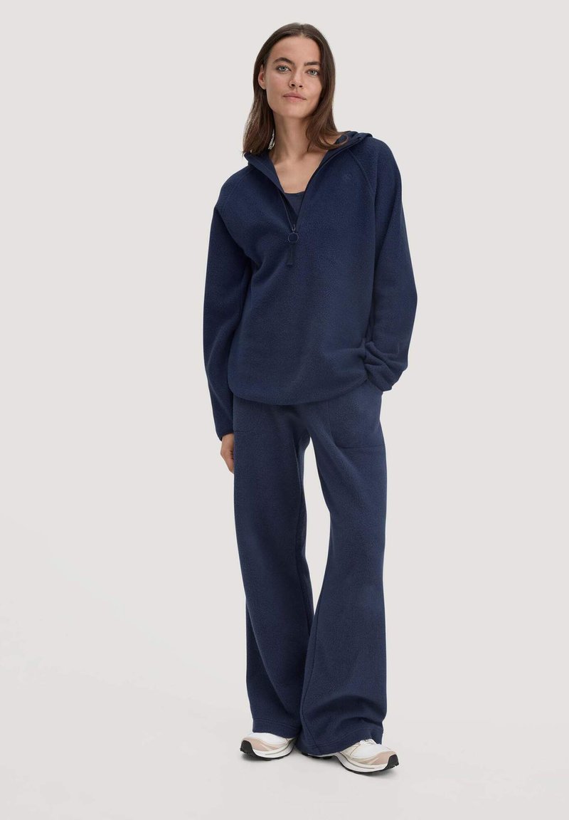 Vrouw die staat in een marineblauwe ritssluitingen fleece sweatshirt en bijpassende wijde fleece broeken met witte sneakers op een lichte achtergrond.