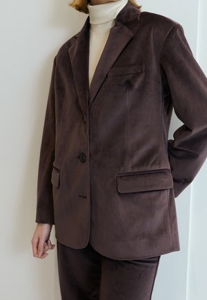 Osoba ubrana w ciemnobrązową zamszową marynarkę z klapami na kieszeniach, w kremowym golfie, stojąca na tle białej ściany.