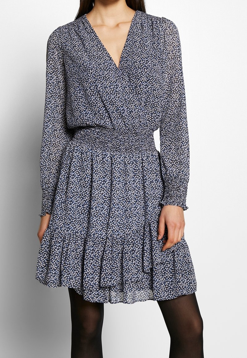 Femme portant une robe portefeuille à manches longues, bleu marine et blanche à motif floral, avec une taille smockée et une jupe plissée, associée à des collants noirs.