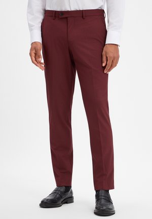 Man die op maat gemaakte bordeauxrode broek, zwarte nette schoenen en een wit overhemd met lange mouwen draagt, staand tegen een effen achtergrond.