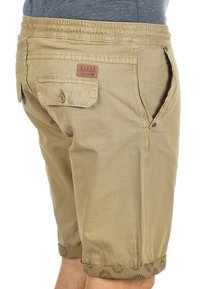 Pantalones cortos de algodón color beige con cintura elástica, bolsillos laterales y un bolsillo trasero con una etiqueta. Decorados con patrones de sol radiante en negro en el dobladillo.