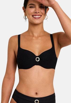 Femme souriante aux yeux fermés portant un bikini noir avec des détails de boucles carrées argentées sur le haut et le bas, sur un fond uni.